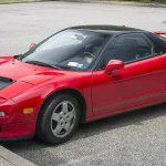 A 1991 Acura NSX in red with the black roof panel of the early coupes, showing the pop-up headlights and low cowl