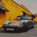 The Porsche 911 Engine Number Is the Originality Detail That Moves the Money Air-cooled Porsche 911 Carrera parked beside a yellow farm building, front three-quarter view