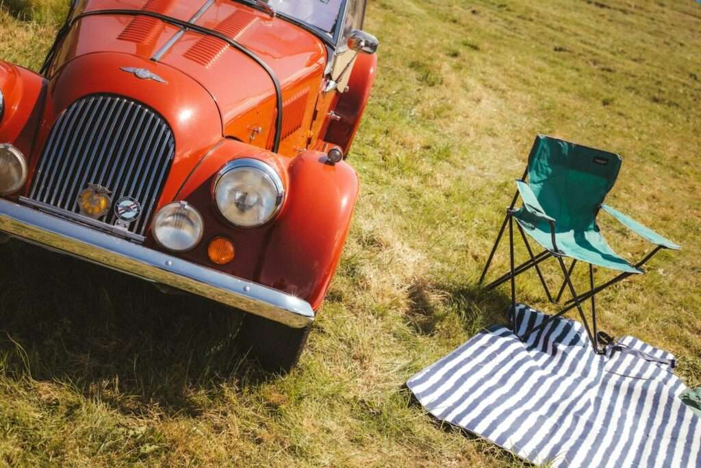 A vintage orange Morgan sports car beside a green folding chair and striped blanket