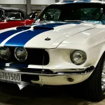 1967 Shelby GT500 fastback in white with blue Le Mans stripes, front three-quarter view at a car show