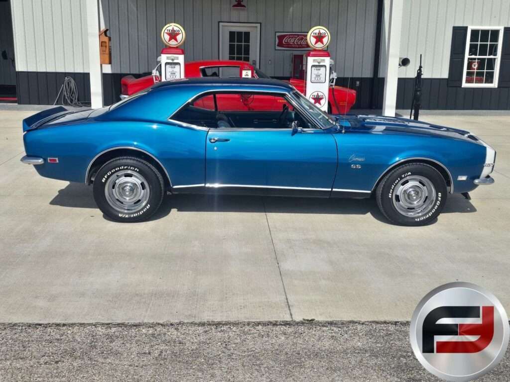Driver-side profile of the blue 1968 Camaro SS parked in front of a garage with vintage gas pumps