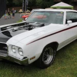 Apollo White 1970 Buick GSX coupe with black hood and stripe treatment parked on grass at a car show