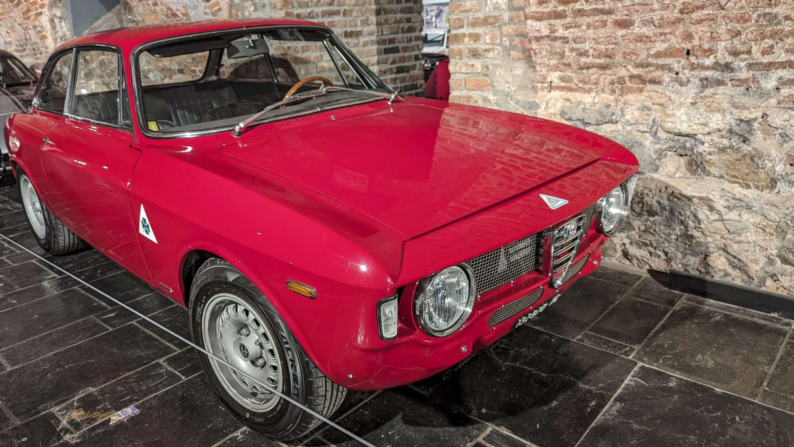 Red 1965 Alfa Romeo Giulia Sprint GTA coupe with steel wheels displayed in a brick-walled museum room