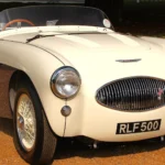 On an Austin-Healey 100S, Alloy Panels and Disc Brakes Prove Little. Recreations Have Both. White 1955 Austin-Healey 100S with dark blue lower sides, small oval grille and low windscreen, seen from the front at a concours