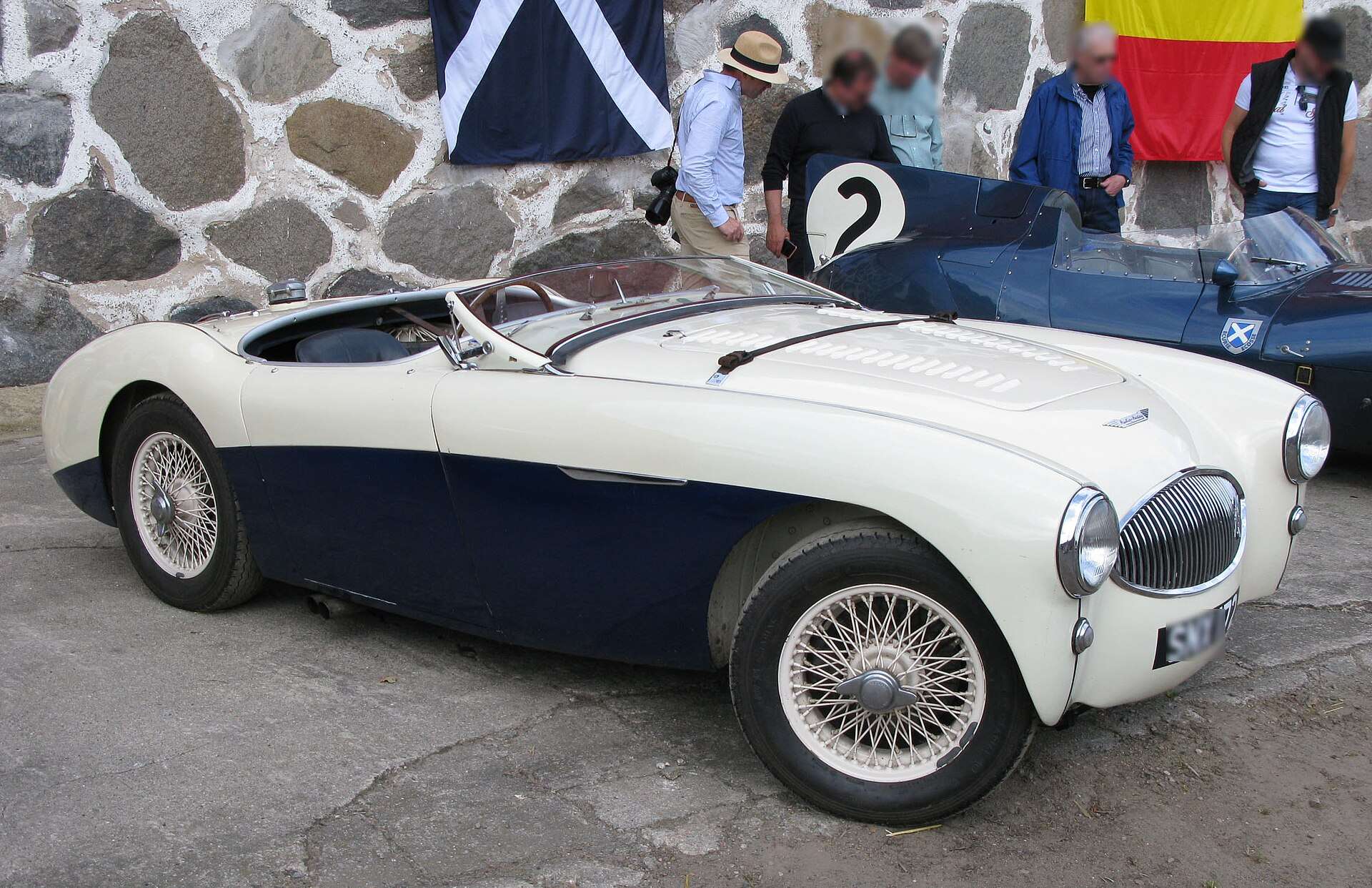 White and dark blue Austin-Healey 100S race car with wire wheels parked beside a stone wall at the Kristianstad Swedish Grand Prix anniversary