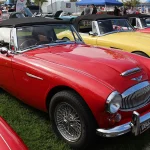 Austin-Healey 3000 vs Sunbeam Tiger vs Triumph TR6: Three British Roadsters, Priced by Condition, Not Badge Red Austin-Healey 3000 Mk III BJ8 roadster with wire wheels and chrome grille parked on grass at a British car show