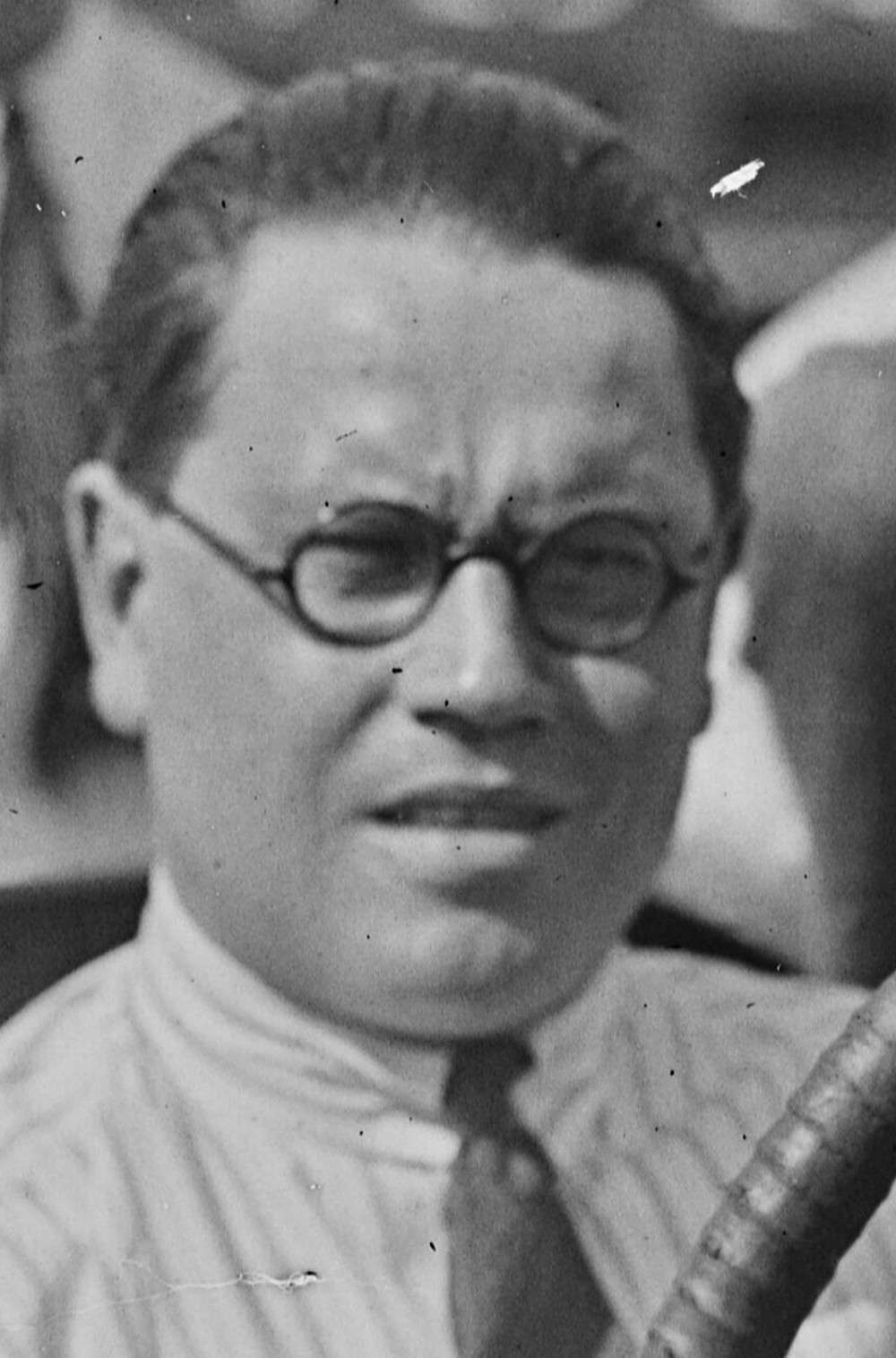 Close black-and-white portrait of racing driver Ernest Eldridge in round spectacles and a striped shirt at the wheel of a race car