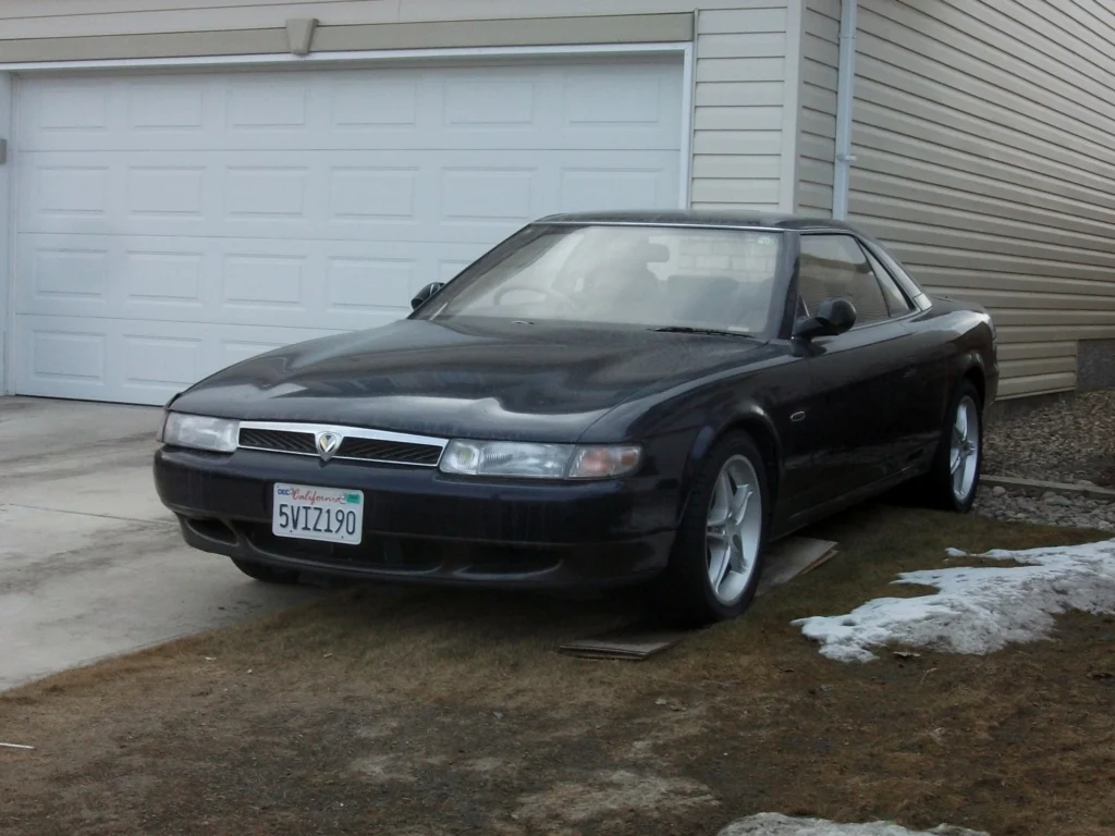 Black Eunos Cosmo coupe with alloy wheels parked in a residential driveway, front three-quarter view