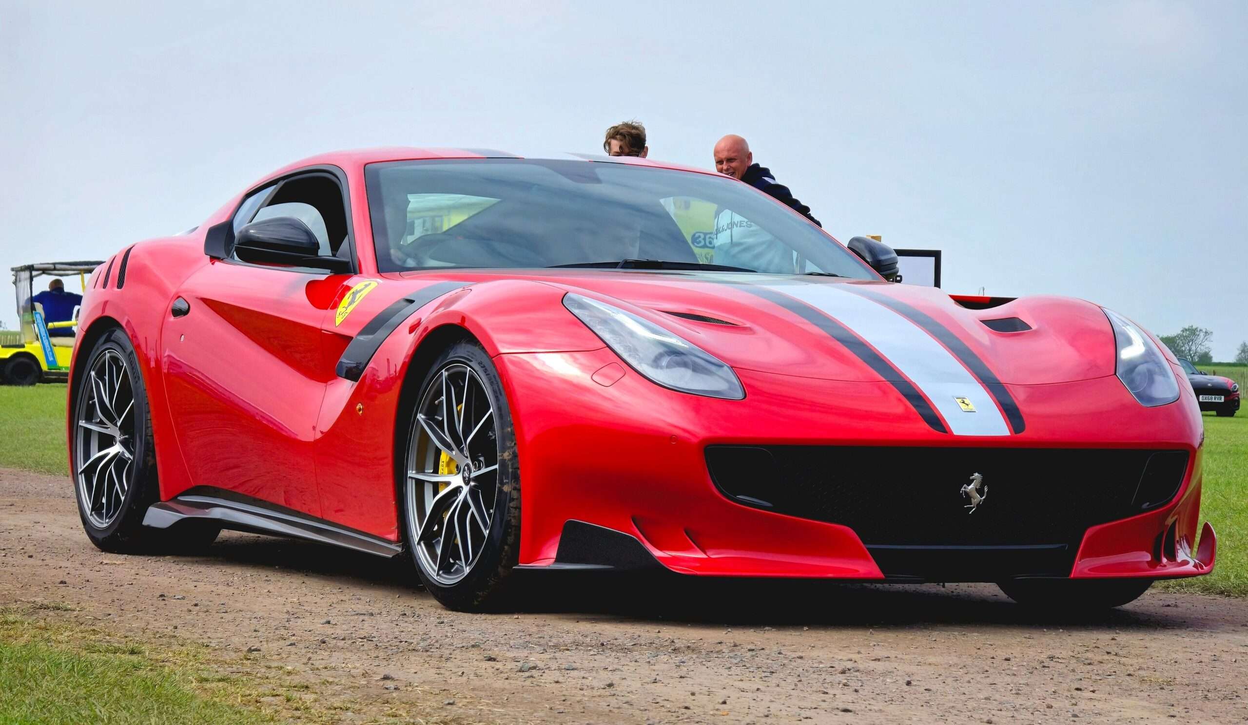 Red Ferrari F12tdf with a black and white hood stripe on a gravel paddock, front three-quarter view