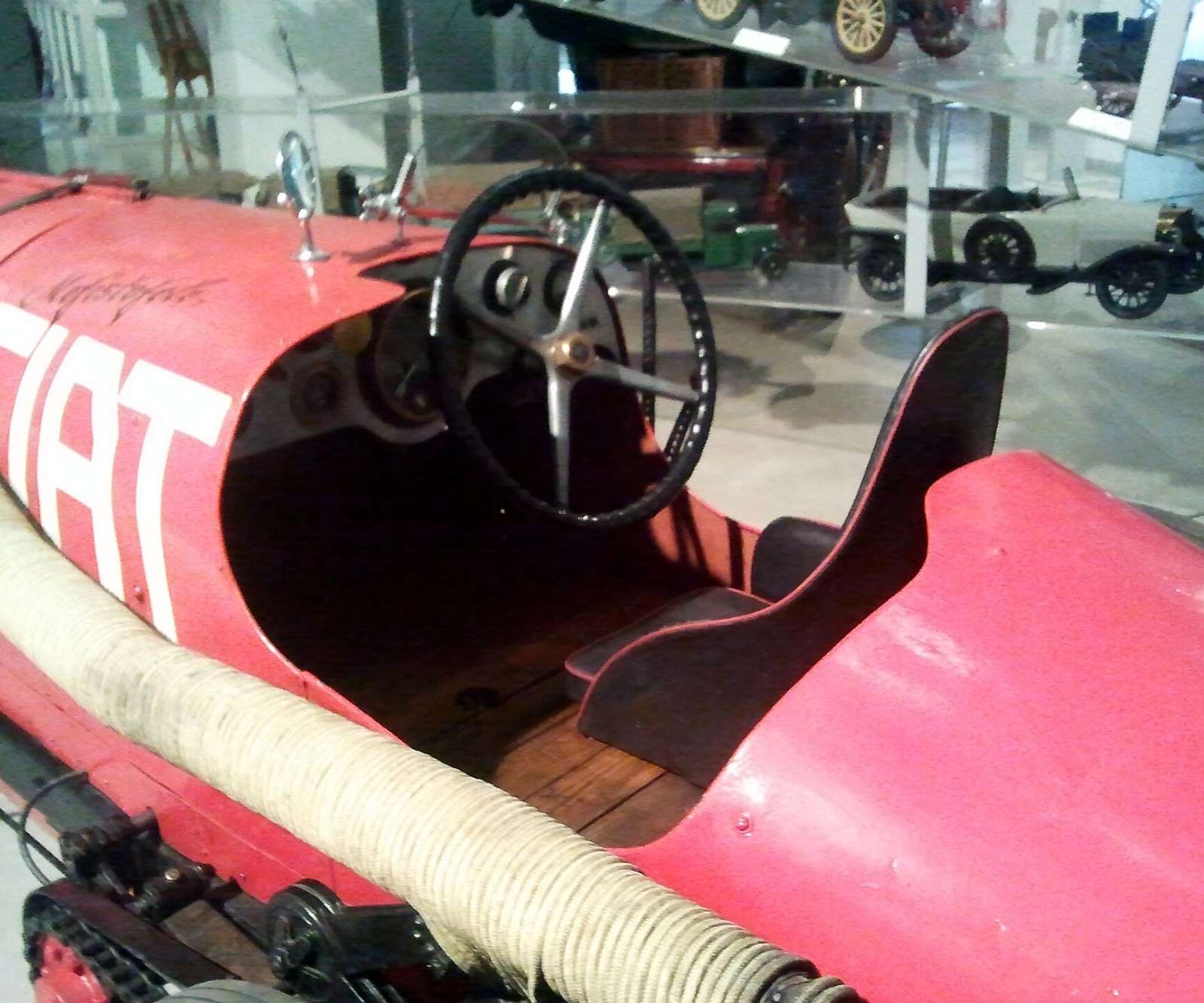 Cockpit of the red Fiat Mefistofele record car in a museum, showing the thin steering wheel, bench seat and large FIAT lettering on the bodywork