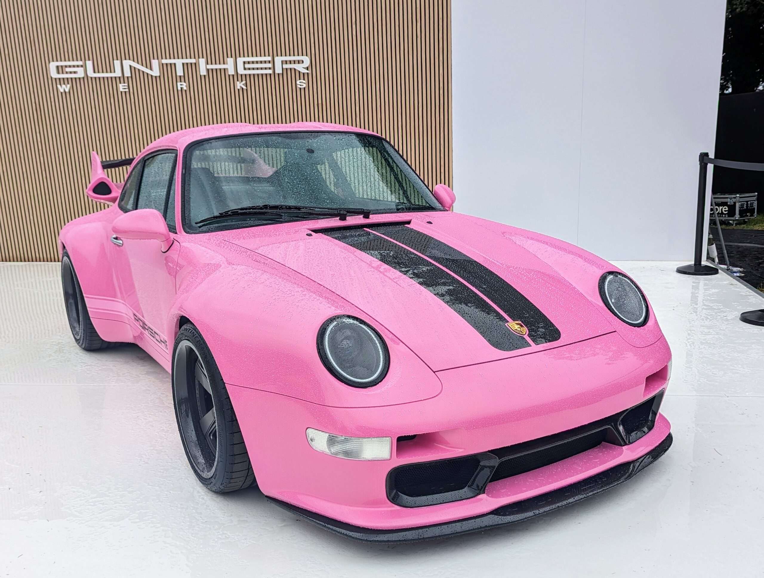 Pink Gunther Werks-modified Porsche 911 with black hood stripes parked in front of a Gunther Werks display wall
