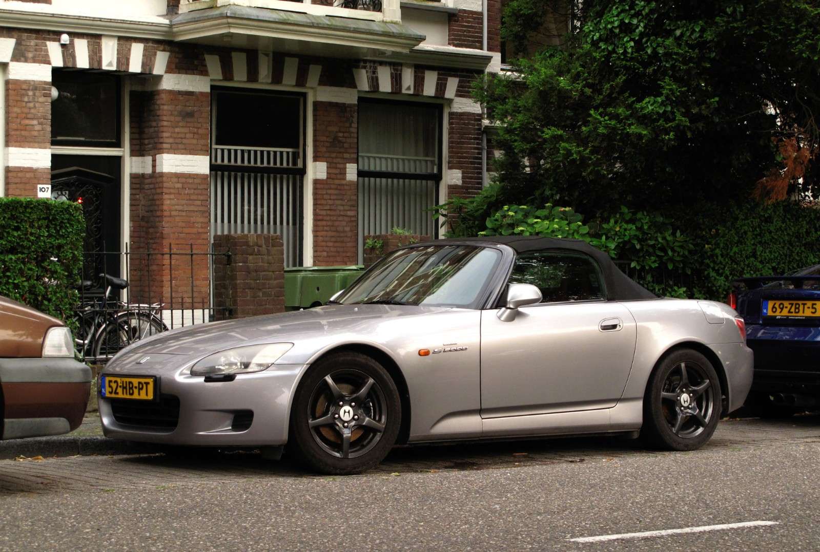Silver first-generation Honda S2000 roadster with its black soft top raised, parked on a tree-lined street in Leiden