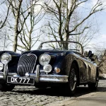 Dark blue Jaguar XK150 with its top down parked on a cobbled street