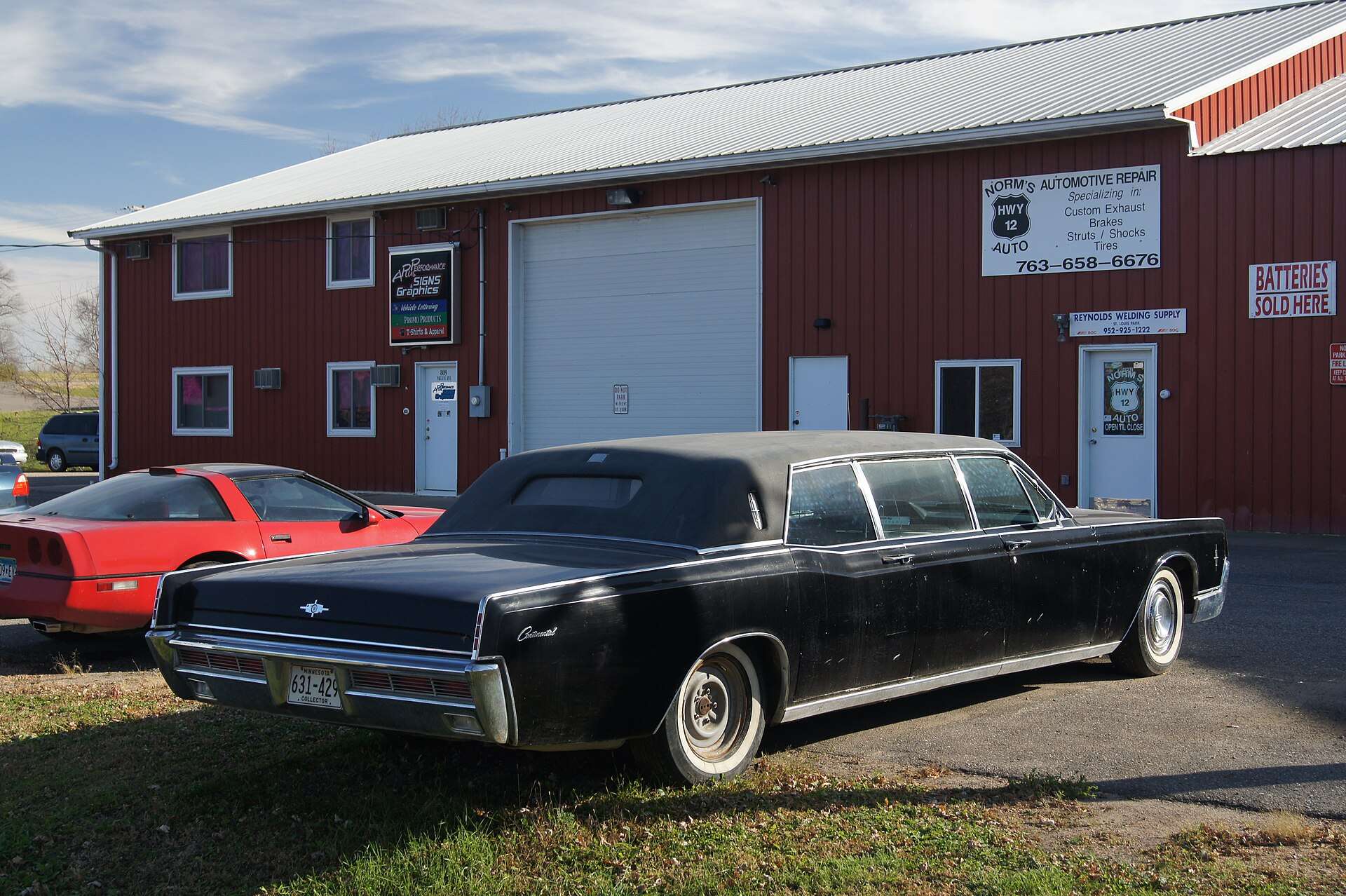 Rear three-quarter view of a black stretched Lincoln Continental limousine with a padded vinyl roof beside a red garage building