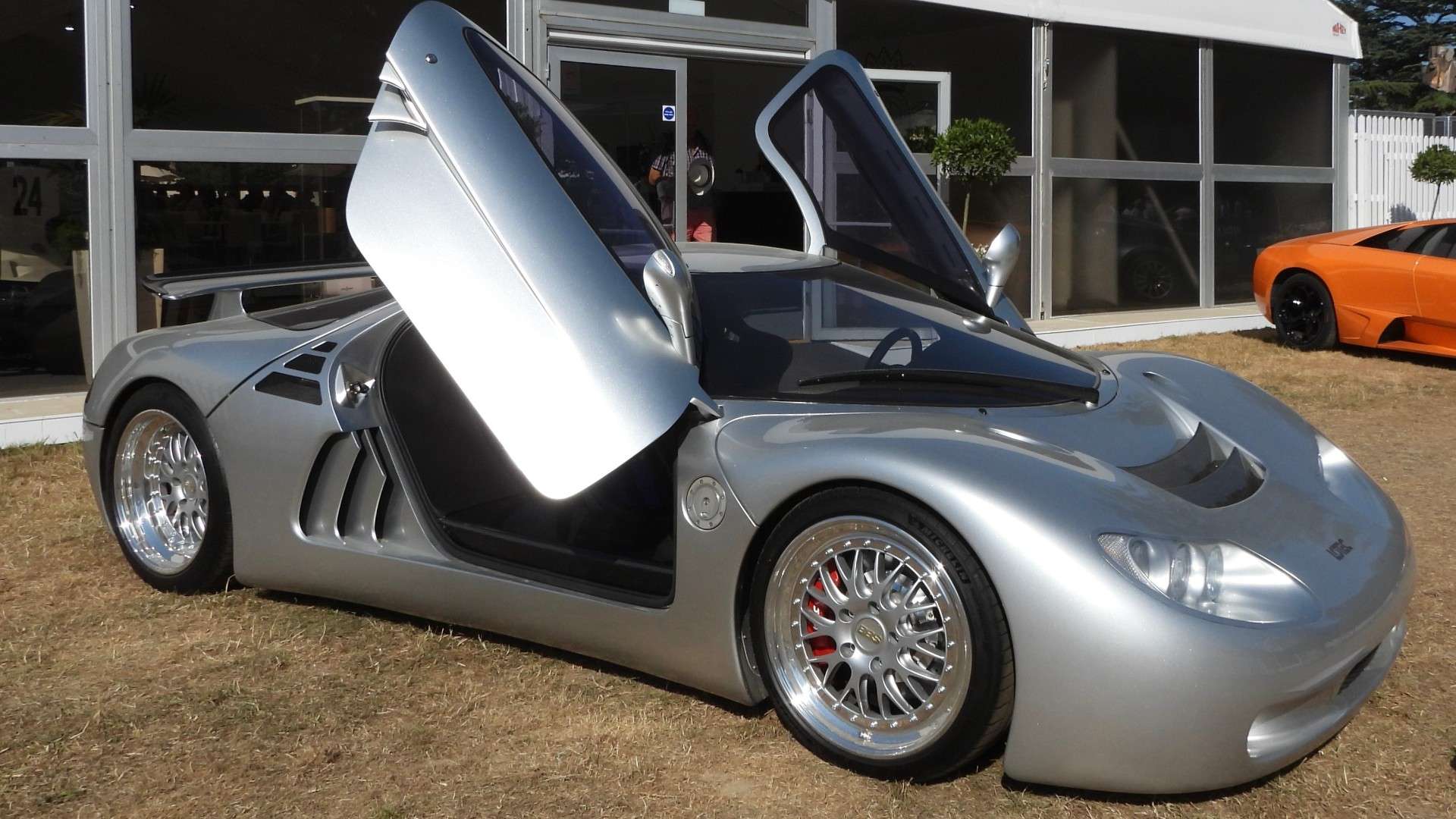 Silver Lotec Sirius supercar with its gullwing-style doors raised, parked on grass at a car show