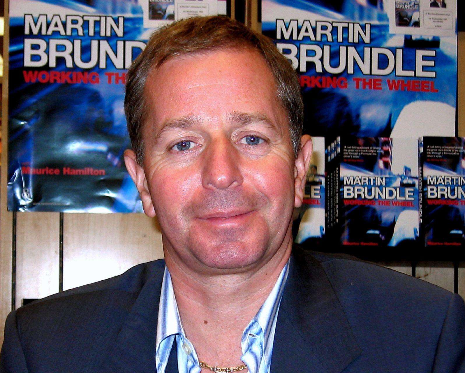 Portrait of Martin Brundle, the former Formula 1 and Le Mans-winning driver, smiling in front of posters for his book Working the Wheel
