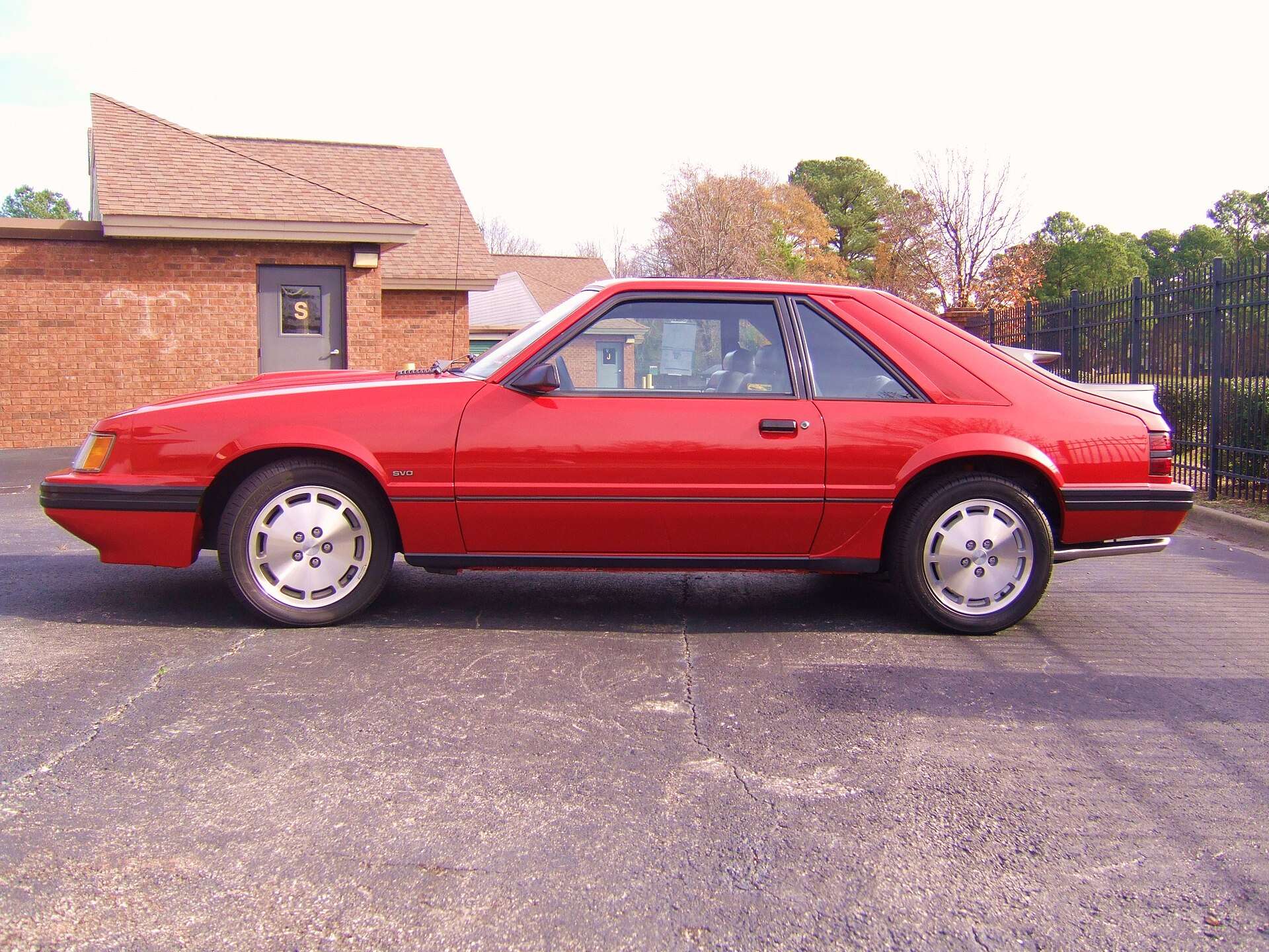 Side profile of a red 1986 Ford Mustang SVO hatchback on 16-inch aluminum wheels