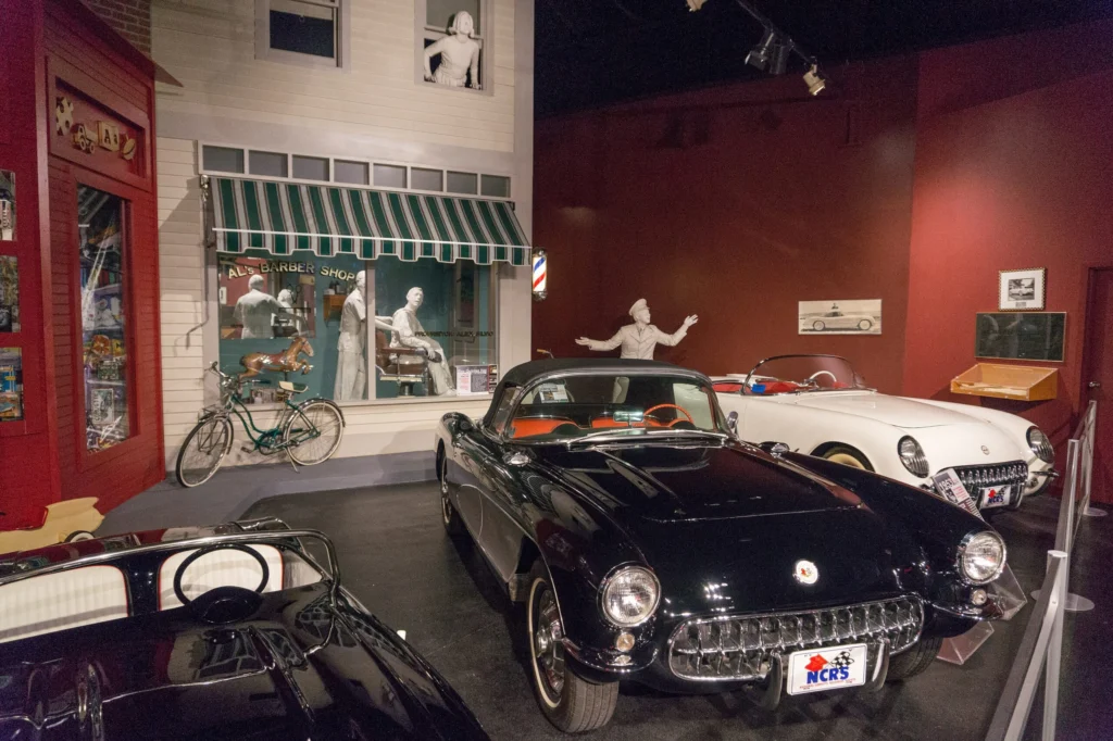 Two early solid-axle Corvettes, one black and one white, parked in a recreated 1950s street scene with a storefront at the National Corvette Museum