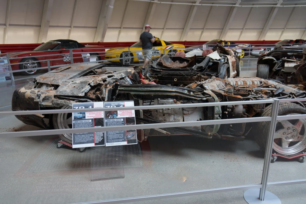 Several heavily damaged, dirt-caked Corvettes displayed behind a barrier with interpretive placards at the National Corvette Museum