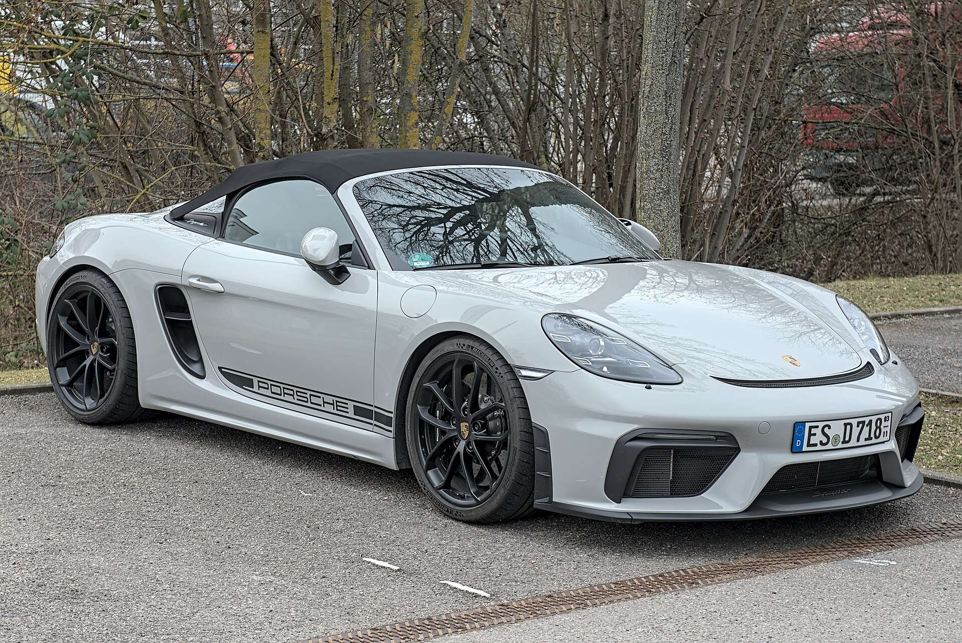 Light gray Porsche 718 Boxster Spyder with black wheels and a black soft top parked on a paved street