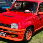 On a Renault 5 Turbo, Aluminum Panels Make the Turbo 1. A Chassis Suffix Alone Won’t Prove It. Red 1981 Renault 5 Turbo 1 with orange bumpers and wheel centers, seen from the front three-quarter at a car show