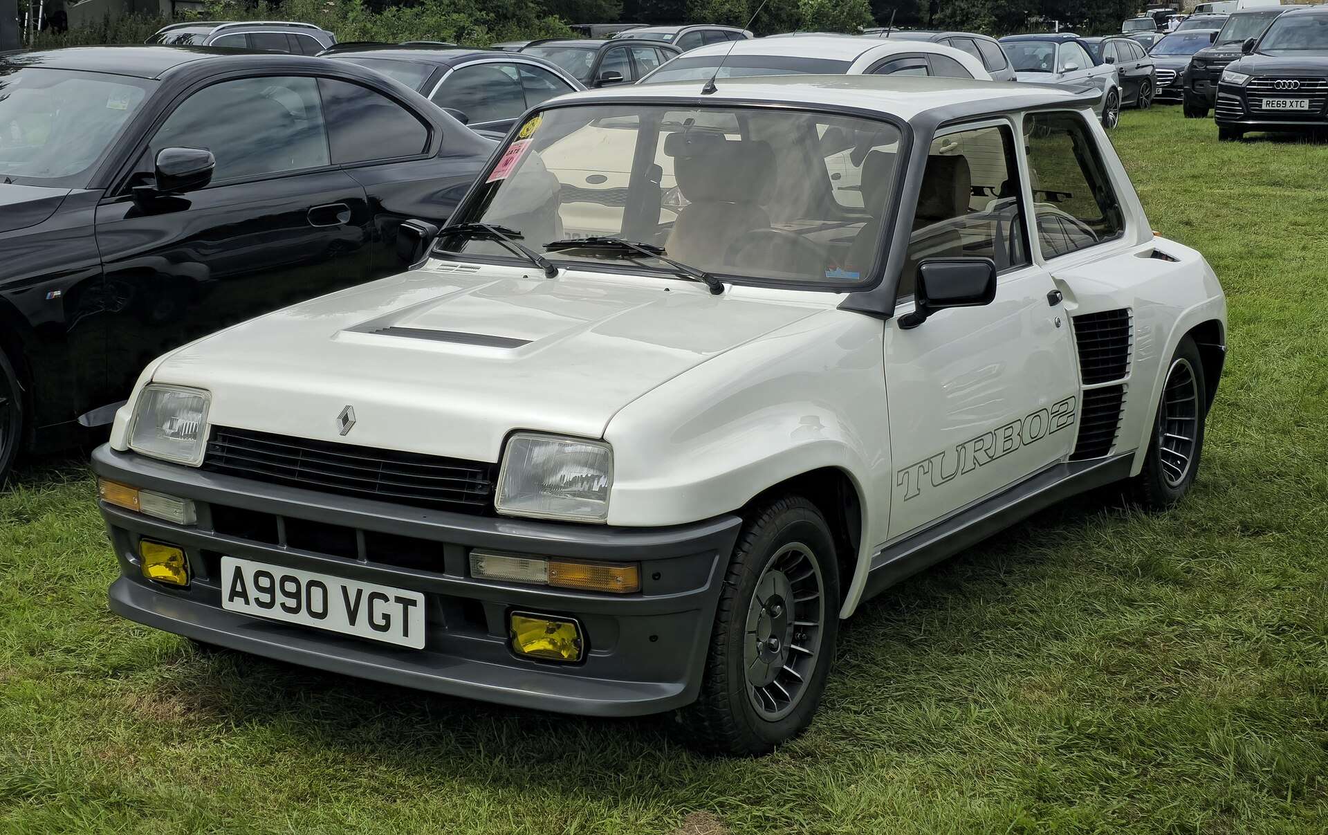 White 1984 Renault 5 Turbo 2 with grey bumpers and black wheels parked on grass, seen from the front three-quarter