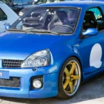 Front three-quarter view of a blue Renault Clio V6 Phase 2 with gold wheels, widened bodywork and a mesh grille
