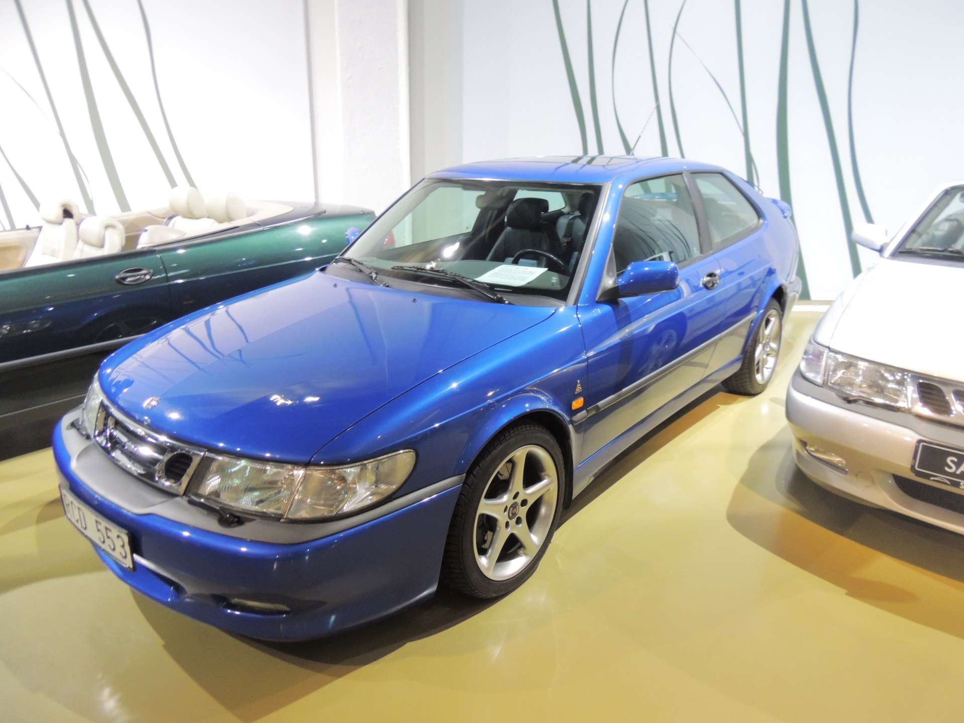 Blue Saab 9-3 Viggen coupe on display indoors with silver and green Saab models beside it