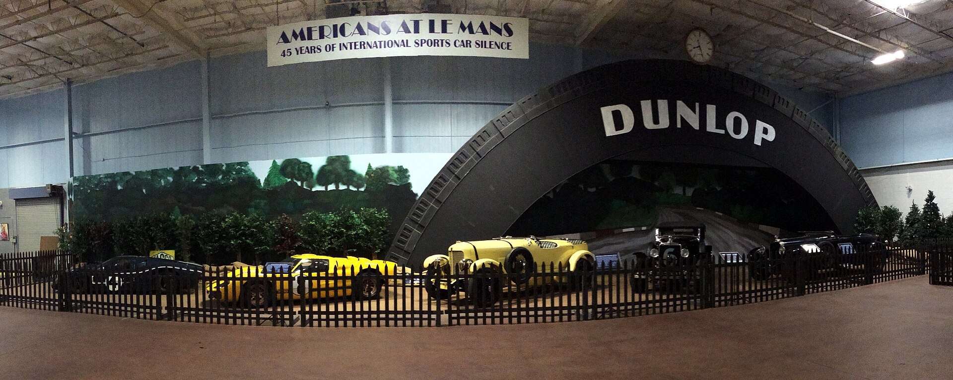 Panoramic view of the Le Mans exhibit at the Simeone Foundation Automotive Museum with a Dunlop Bridge backdrop and racing cars in front