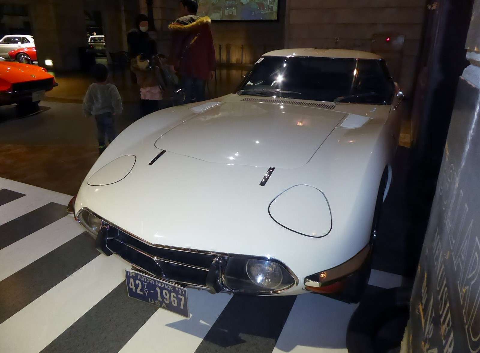 White Toyota 2000GT coupe photographed from the front at a museum, showing its long hood and recessed headlamps