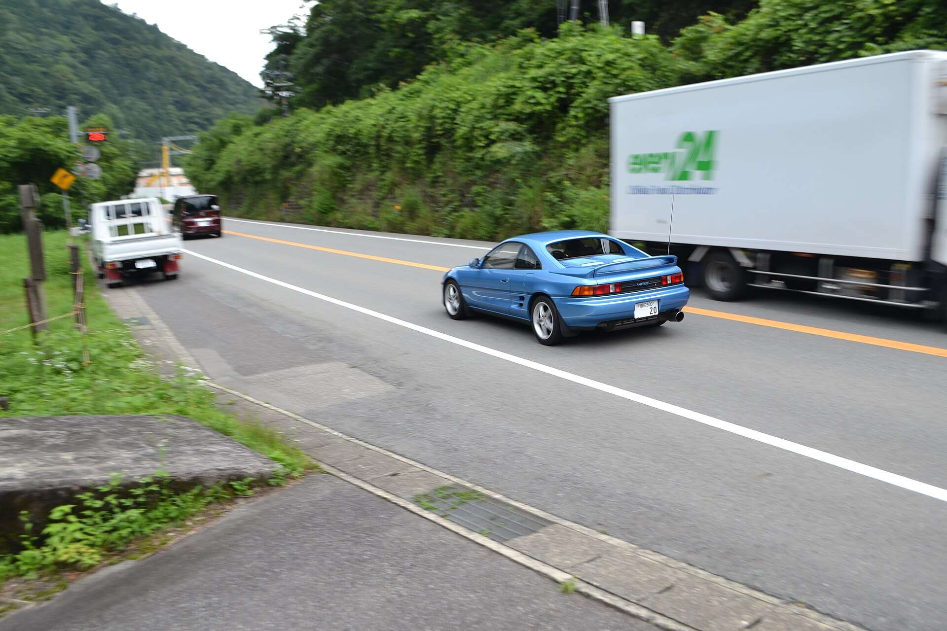 Blue second-generation Toyota MR2 SW20 coupe driving on a two-lane road past a truck