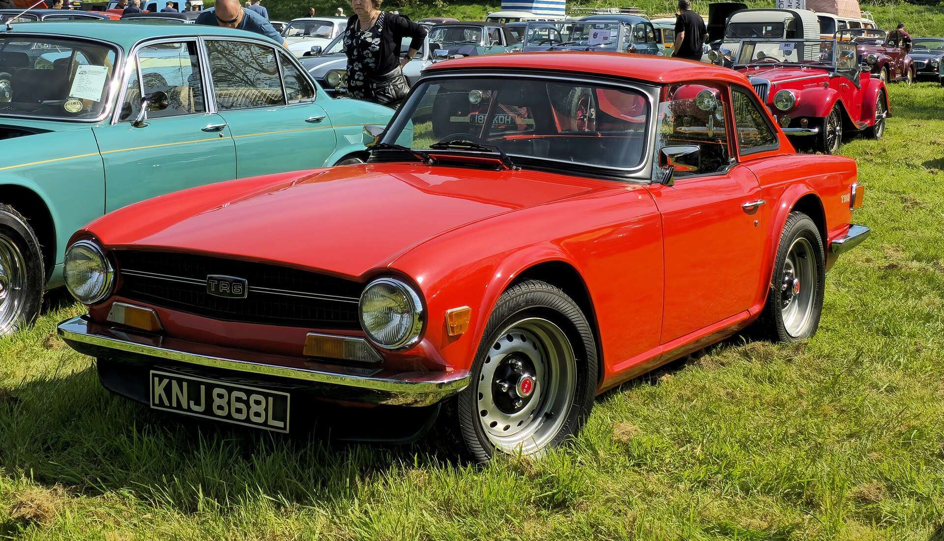 Red 1972 Triumph TR6 roadster with black grille and chrome bumper at an outdoor classic car show