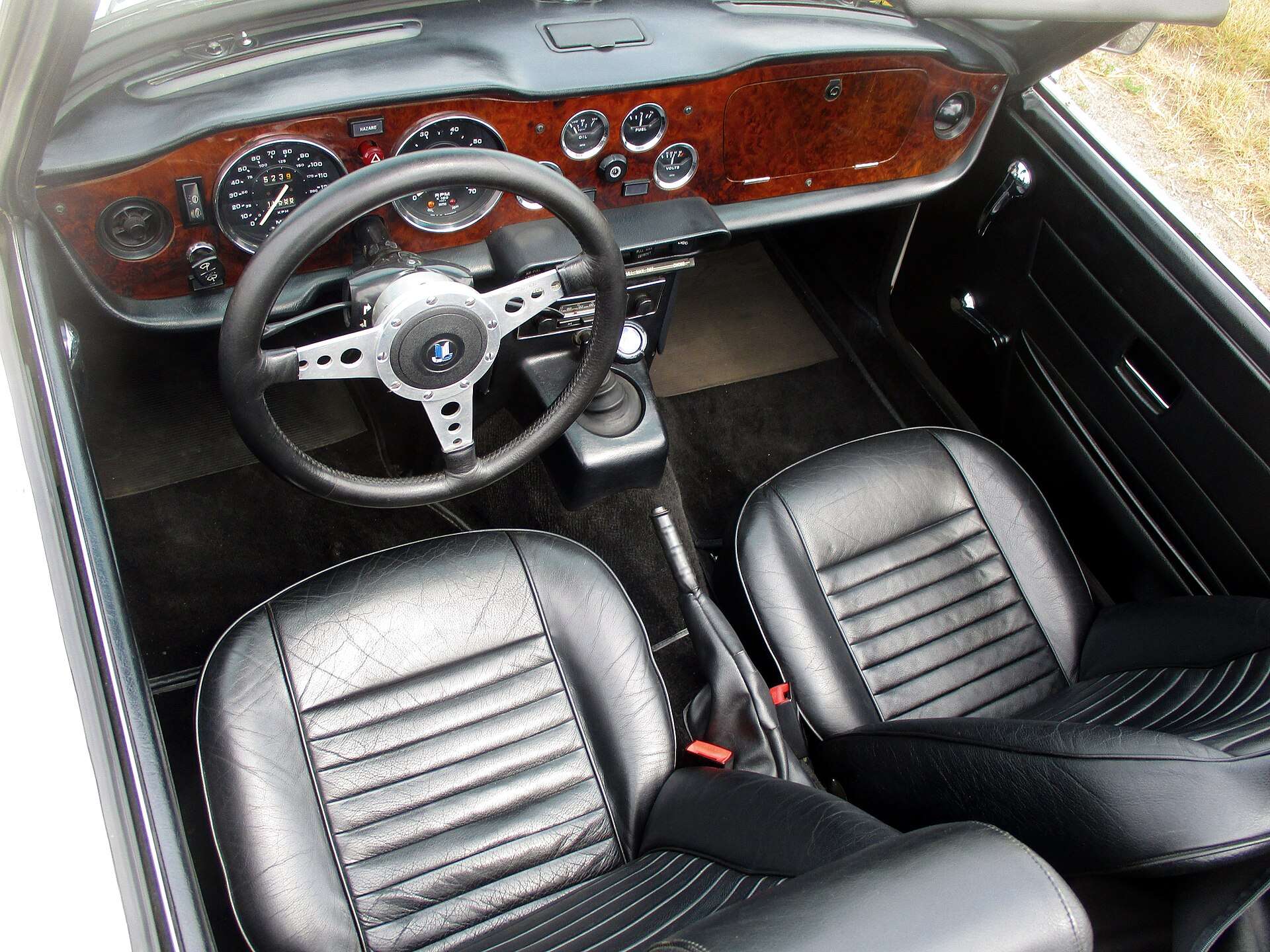 Interior of a Triumph TR6 showing wood dashboard, wood-rim steering wheel and black leather bucket seats