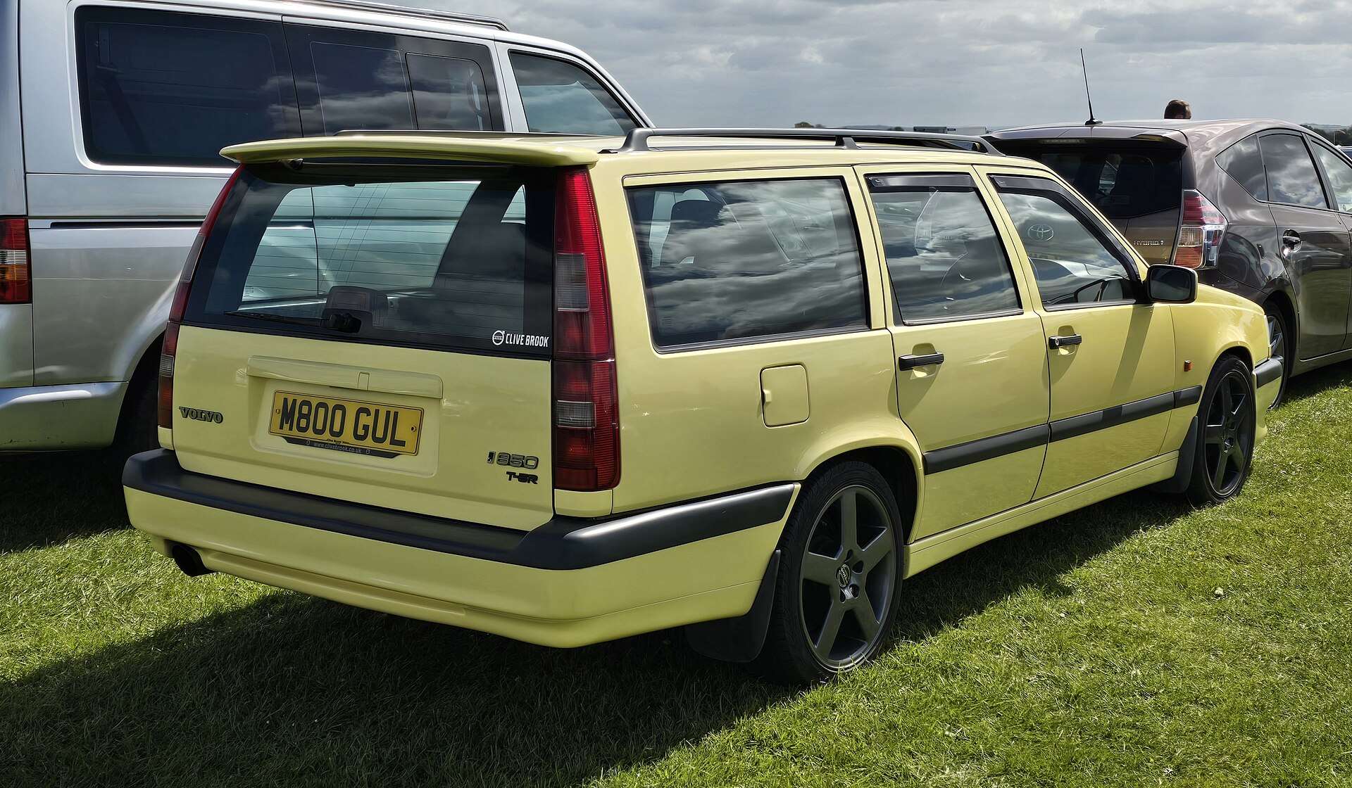 Rear three-quarter view of a Cream Yellow Volvo 850 T-5R estate with roof rails and a rear spoiler, parked on grass